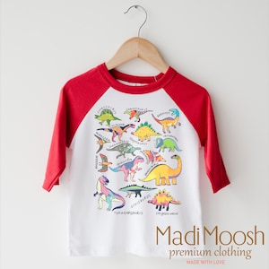 May include: A white and red long-sleeved t-shirt with a colorful dinosaur graphic print. The shirt has a red baseball-style sleeve design. The text "Madi Moosh premium clothing MADE WITH LOVE" is printed on the bottom of the shirt.