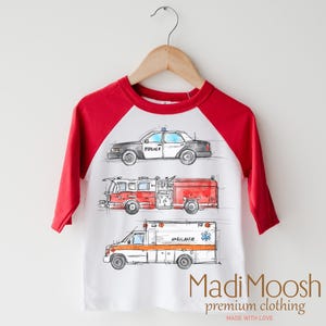 May include: A white and red baseball tee shirt with a graphic of a police car, fire truck, and ambulance. The text "Madi Moosh premium clothing MADE WITH LOVE" is printed on the bottom of the shirt.