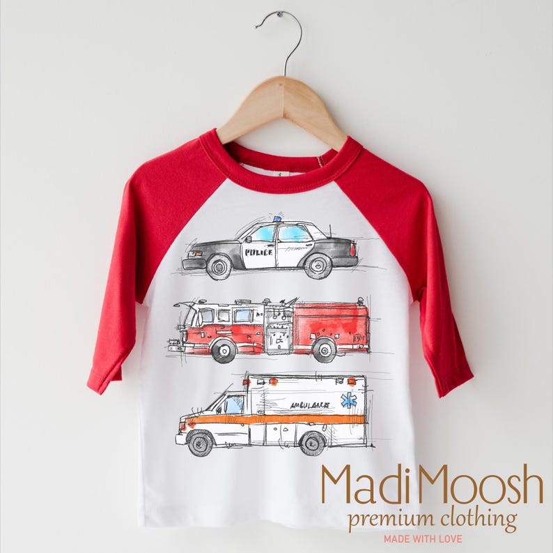 May include: A white and red baseball tee shirt with a graphic of a police car, fire truck, and ambulance. The text "Madi Moosh premium clothing MADE WITH LOVE" is printed on the bottom of the shirt.