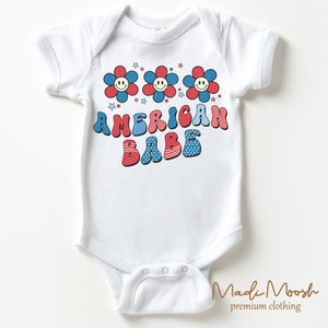 May include: White baby bodysuit with a retro design. The bodysuit features three smiling flower graphics in red and blue, with the text "AMERICAN BABE" in a matching color scheme. Stars are scattered above the text.