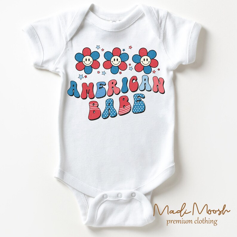 May include: White baby bodysuit with a retro design. The bodysuit features three smiling flower graphics in red and blue, with the text "AMERICAN BABE" in a matching color scheme. Stars are scattered above the text.