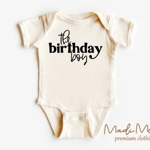 May include: Cream-colored baby bodysuit with the text "the birthday boy" in black script. The bodysuit is made of soft material and has short sleeves. The brand name "Madi Moosh" is printed at the bottom.