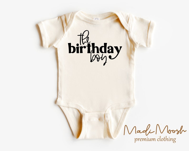 May include: Cream-colored baby bodysuit with the text "the birthday boy" in black script. The bodysuit is made of soft material and has short sleeves. The brand name "Madi Moosh" is printed at the bottom.