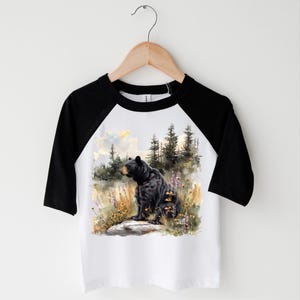 May include: A white and black raglan baseball tee featuring a watercolor illustration of a black bear sitting in a field of wildflowers and tall grass. The bear is facing left, with a forest and sky in the background.