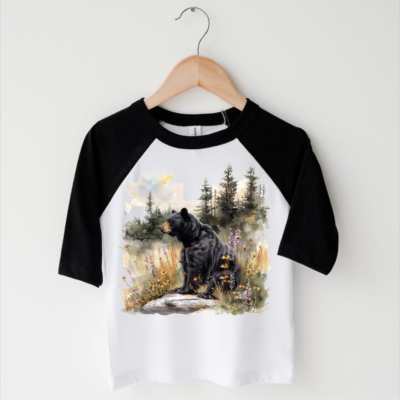 May include: A white and black raglan baseball tee featuring a watercolor illustration of a black bear sitting in a field of wildflowers and tall grass. The bear is facing left, with a forest and sky in the background.