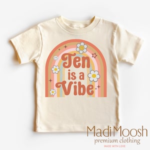 May include: A cream colored t-shirt with a rainbow design and the text "Ten is a Vibe" in orange and brown letters. The rainbow has white daisies and stars. The text "Madi Moosh premium clothing MADE WITH LOVE" is at the bottom of the shirt.