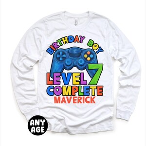 May include: White long-sleeve t-shirt with a colorful graphic of a video game controller and the text "Birthday Boy Level 7 Complete Maverick".