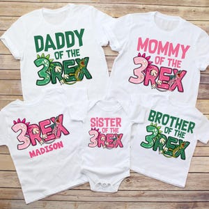 May include: Set of five white t-shirts with green and pink dinosaur graphics. The shirts say "Daddy of the 3Rex", "Mommy of the 3Rex", "Sister of the 3Rex", "Brother of the 3Rex", and "Madison" with a pink dinosaur graphic.