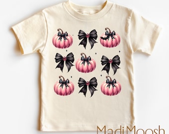 May include: A cream-coloured t-shirt with a pattern of pink pumpkins and black bows. The pumpkins are arranged in a grid, with black bows alternating with the pumpkins. The text "MadiMoosh premium clothing MADE WITH LOVE" is printed at the bottom.