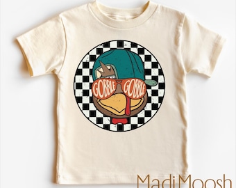 May include: Cream-colored t-shirt featuring a cartoon turkey wearing sunglasses that read "GOBBLE GOBBLE" inside a black and white checkered circle. The turkey is wearing a teal baseball cap and a red bow tie. The shirt has the text "MadiMoosh premium clothing" at the bottom.