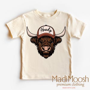 May include: A cream colored t-shirt with a cartoon illustration of a brown Highland cow wearing a red baseball cap with the word "Howdy" on it.