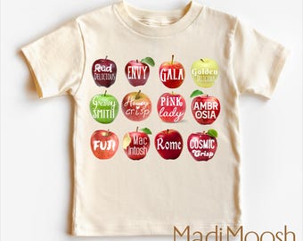 May include: Cream-colored t-shirt featuring illustrations of various apple varieties, each labeled with its name in white script. The shirt includes the text "Madi Moosh premium clothing MADE WITH LOVE" at the bottom.