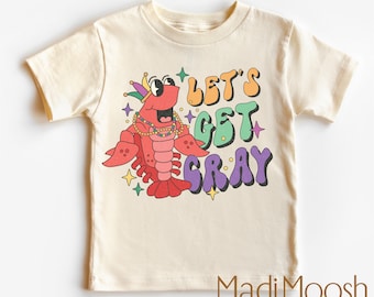 May include: Cream-coloured toddler t-shirt with a cartoon lobster wearing a Mardi Gras hat and necklace. The shirt has the text "LET'S GET CRAY" in colourful, retro-style lettering. The brand name "Madi Moosh" is printed at the bottom.