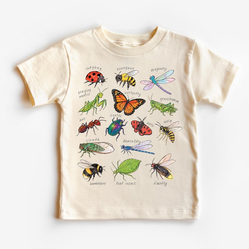 May include: A cream-coloured t-shirt displaying colourful illustrations of insects such as a ladybug, butterfly, and dragonfly. Each insect is labelled with its name in a handwritten font. The shirt is made of a soft material.