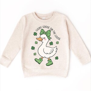 May include: Beige sweatshirt featuring a cartoon goose with a green bow, boots, and shamrocks. The text "LUCKY GOOSE ON THE LOOSE" arches above the goose. A St. Patrick's Day themed design.