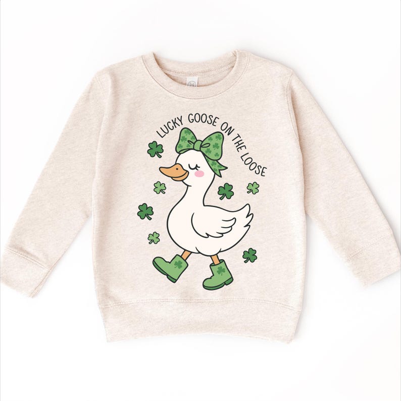May include: Beige sweatshirt featuring a cartoon goose with a green bow, boots, and shamrocks. The text "LUCKY GOOSE ON THE LOOSE" arches above the goose. A St. Patrick's Day themed design.