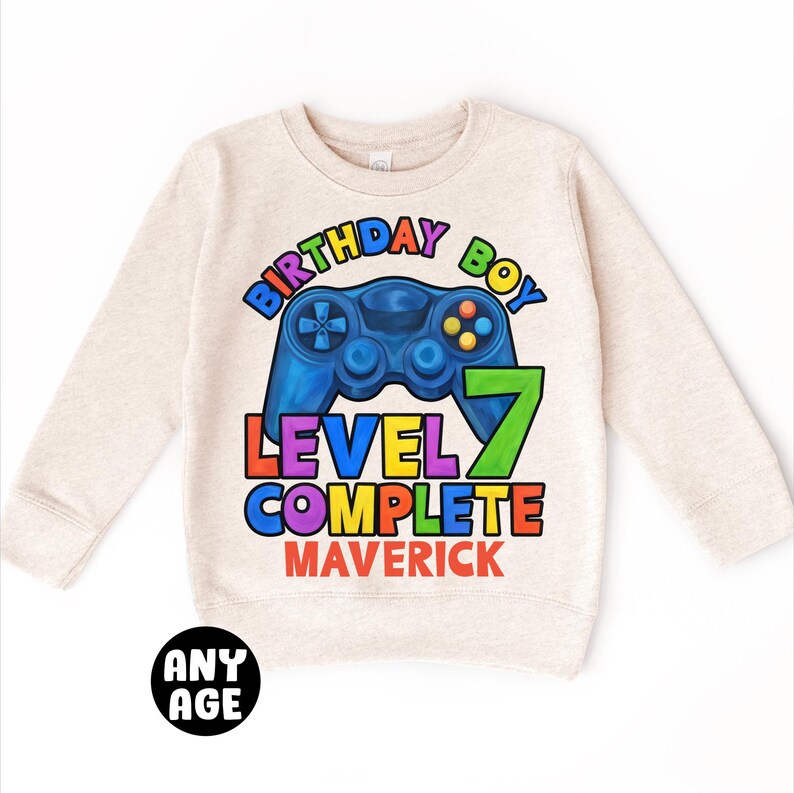 May include: A beige sweatshirt with a colorful graphic of a video game controller and the text "Birthday Boy Level 7 Complete Maverick".
