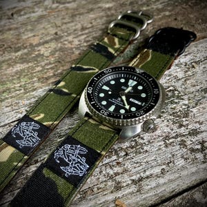 May include: A black Seiko dive watch with a stainless steel bezel and a black dial. The watch is paired with a green and brown camouflage patterned watch strap. The strap features a white embroidered emblem.