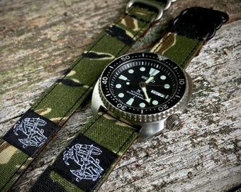 May include: A black Seiko dive watch with a stainless steel bezel and a black dial. The watch is paired with a green and brown camouflage patterned watch strap. The strap features a white embroidered emblem.