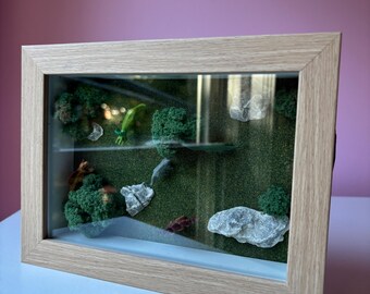 May include: A framed miniature diorama with a natural landscape scene. The scene includes green moss, small trees, rocks, and a small green lizard. The frame is made of light-colored wood, and the background is a pink wall.
