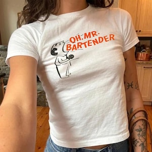 May include: White t-shirt with the text "OH MR. BARTENDER" in orange and a black and white cartoon illustration of a bartender. The shirt is short-sleeved and has a crew neck.