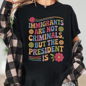 May include: Black t-shirt with the text "IMMIGRANTS ARE NOT CRIMINALS, BUT THE PRESIDENT IS" in colorful, retro-style lettering. The design includes flower accents and rainbow stripes, creating a bold statement.