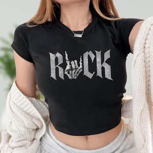 A woman wearing a black crop top with the word "ROCK" printed on it, along with a skeleton hand graphic. She is also wearing a necklace and has a white sweater draped over her shoulders.