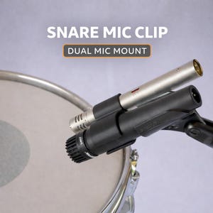 Könnte beinhalten: Eine schwarze Doppel-Mikrofonhalterung mit zwei Mikrofonen, die über einer Snare-Drum positioniert sind. Das obere Mikrofon ist silberfarben und das untere Mikrofon ist schwarz. Der Text oben lautet "SNARE MIC CLIP" und "DUAL MIC MOUNT".
