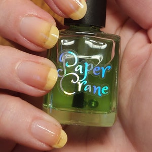 Puede incluir: Una botella de vidrio transparente de esmalte de uñas verde con una etiqueta amarilla que dice "Paper Crane". La botella está siendo sostenida por una mano con esmalte de uñas amarillo en las uñas.