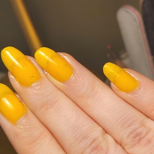 Puede incluir: Primer plano de una mano con uñas largas y ovaladas pintadas de un amarillo vibrante y brillante. Las uñas tienen un acabado liso y uniforme, que refleja la luz. El tono de piel es claro, lo que contrasta con el color brillante de las uñas.
