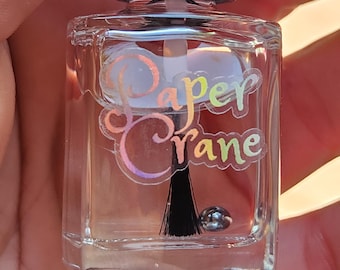 May include: Clear nail polish bottle with a black cap and brush. The bottle has a holographic label with the words "Paper Crane" in a script font. A small, silver bead sits at the bottom of the bottle.