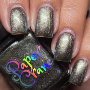 May include: A dark green nail polish with a shimmery finish. The polish is applied to several fingernails, showing the color and texture. The bottle of nail polish is labeled "Paper Frame".