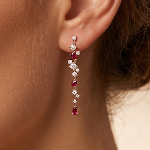 Op de afbeelding: Hangende oorbellen met een delicaat ontwerp. De oorbellen hebben een reeks kleine, sprankelende witte stenen en levendige rode edelstenen. De stenen zijn in een trapsgewijs patroon gerangschikt, wat een elegant accessoire creëert.