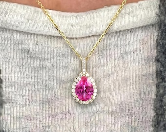 May include: A gold chain necklace featuring a pear-shaped pink gemstone pendant surrounded by a halo of small white stones. The pendant hangs from a delicate gold link chain, resting against a white textured fabric and a gray knit garment.