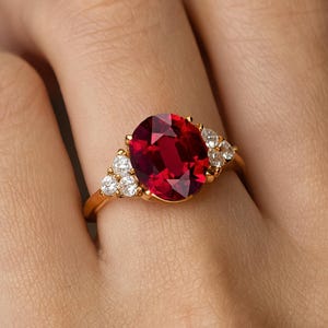 Może przedstawiać: Złoty pierścionek z dużym, owalnym, ciemnoczerwonym kamieniem szlachetnym jako centralnym elementem. Pierścionek jest ozdobiony mniejszymi, przezroczystymi kamieniami szlachetnymi po obu stronach. Pierścionek jest osadzony w złotej obrączce, tworząc klasyczny i elegancki design.