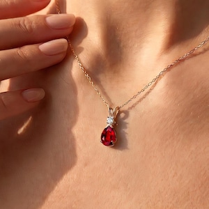 May include: A delicate gold chain necklace featuring a teardrop-shaped red gemstone pendant. The pendant is accented with a single small, clear round gemstone set directly above the red stone, suspended on a thin, polished gold-tone metal chain.
