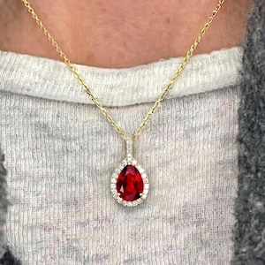 May include: A gold chain necklace featuring a pear-shaped red gemstone pendant surrounded by a halo of small white stones. The pendant hangs from a delicate gold link chain, resting against a white textured fabric and a grey textured garment.