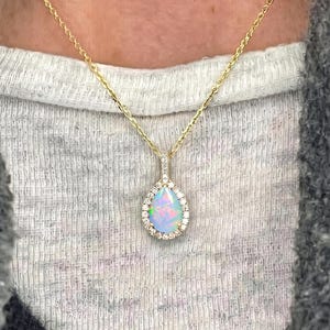 Op de afbeelding: Een gouden ketting met een peervormige opaal hanger omringd door een halo van kleine heldere steentjes. De iriserende opaal vertoont flitsen van blauw, groen en roze, gezet in een verfijnd gouden montuur dat aan een dunne gouden ketting hangt.