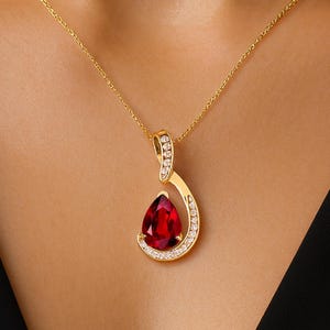 Puede incluir: Un collar con colgante de oro con una piedra preciosa roja oscura en forma de lágrima. El colgante está adornado con pequeñas piedras blancas brillantes y cuelga de una delicada cadena de oro. El collar se muestra sobre un fondo oscuro.