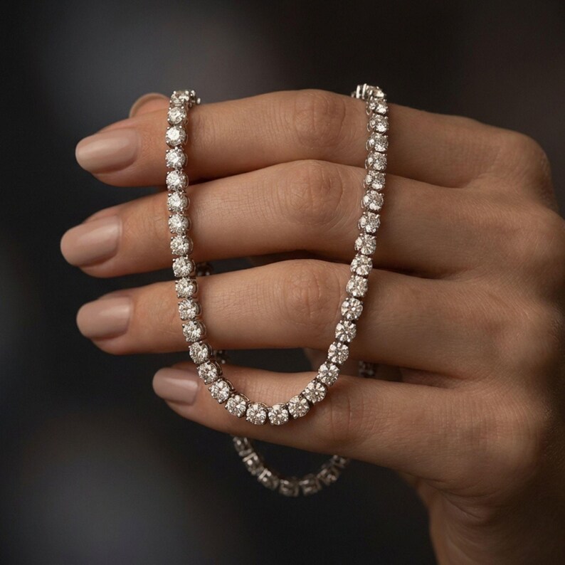 May include: A sparkling diamond tennis necklace. The necklace features a series of round-cut diamonds set closely together, creating a continuous line of brilliance. The diamonds are set in a silver or white gold setting, and the necklace is held in a hand.