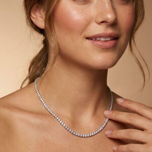May include: A delicate silver necklace with numerous small, round, clear stones. The necklace is worn around the neck and rests on the collarbone. The stones sparkle, reflecting light. The background is a soft, neutral color.