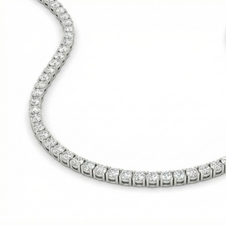 May include: A sparkling, silver-toned tennis necklace. The necklace is composed of numerous round, clear gemstones set closely together. The gemstones are evenly spaced and create a continuous line of brilliance. The background is a clean, bright white.