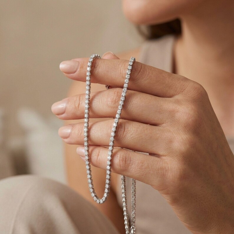 May include: A delicate silver necklace with numerous small, round, clear gemstones. The necklace is held up, showcasing its design and sparkle. The gemstones are evenly spaced along the chain, creating a continuous line of brilliance.