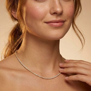 May include: A delicate gold necklace with small, sparkling stones. The necklace is worn around the neck, with the stones catching the light. The jewelry is a classic and elegant accessory.