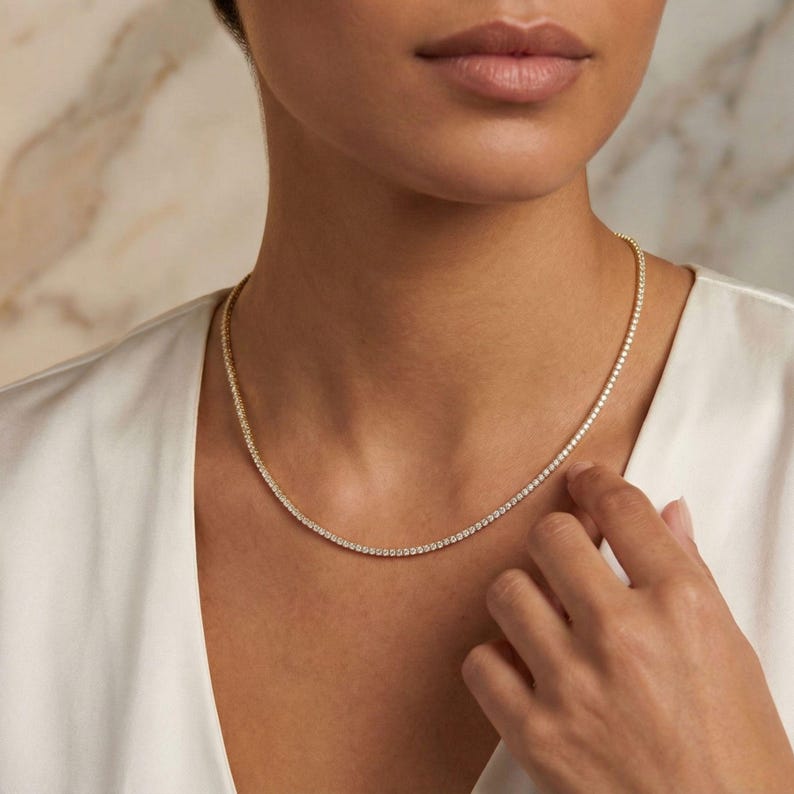 May include: A delicate gold necklace with numerous small, sparkling stones. The necklace is worn around the neck and rests against a white garment. The stones are evenly spaced, creating a continuous line of sparkle.