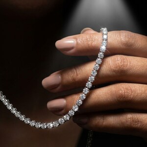 May include: A tennis necklace with numerous round, brilliant-cut diamonds. The diamonds are set closely together in a row, creating a continuous sparkle. The necklace is held against a dark background, highlighting the brilliance of the stones.