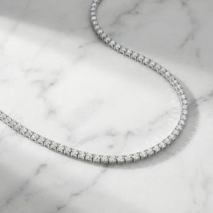 May include: A delicate, silver-toned necklace featuring a continuous line of sparkling, round-cut gemstones. The necklace is displayed on a white marble surface, showcasing its elegant design and brilliance. The gemstones are evenly spaced.