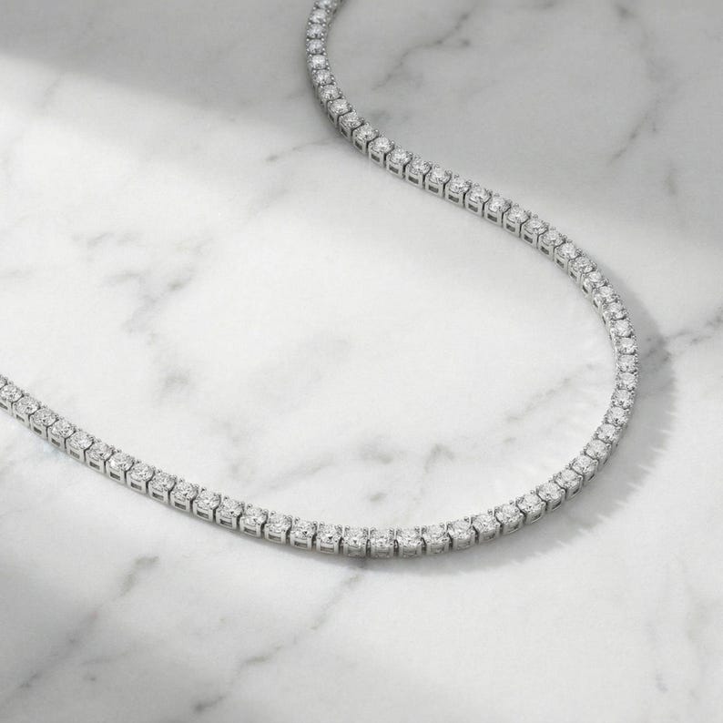 May include: A delicate, silver-toned necklace featuring a continuous line of sparkling, round-cut gemstones. The necklace is displayed on a white marble surface, showcasing its elegant design and brilliance. The gemstones are evenly spaced.