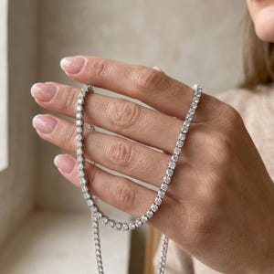 May include: A delicate, silver-toned necklace with numerous small, round, clear gemstones. The necklace is held in a hand with manicured nails, showcasing the jewelry's sparkle and design. The gemstones are evenly spaced along the chain.