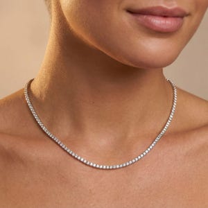 May include: A delicate, silver-toned necklace with numerous small, sparkling stones. The necklace is worn around the neck, resting against the skin. The stones are evenly spaced, creating a continuous line of brilliance. The background is a soft, neutral tone.